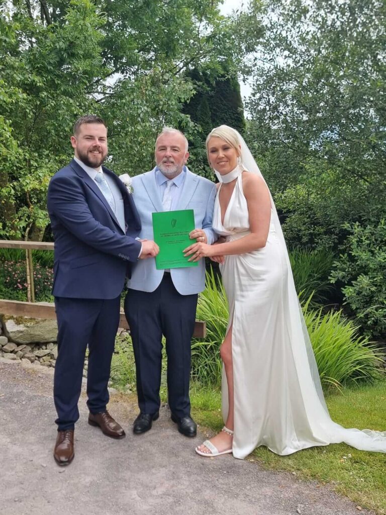 Bride and groom holding their marriage certificate with their celebrant in an outdoor setting.