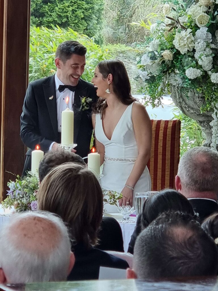 Bride and groom smiling at each other while lighting a unity candle during their wedding ceremony at Muckross Park Hotel surrounded by floral arrangements.