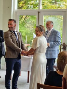 Bride and groom holding hands and smiling during their wedding ceremony at Seaview House with the celebrant Brian standing behind them in a room with large windows and a green outdoor backdrop.