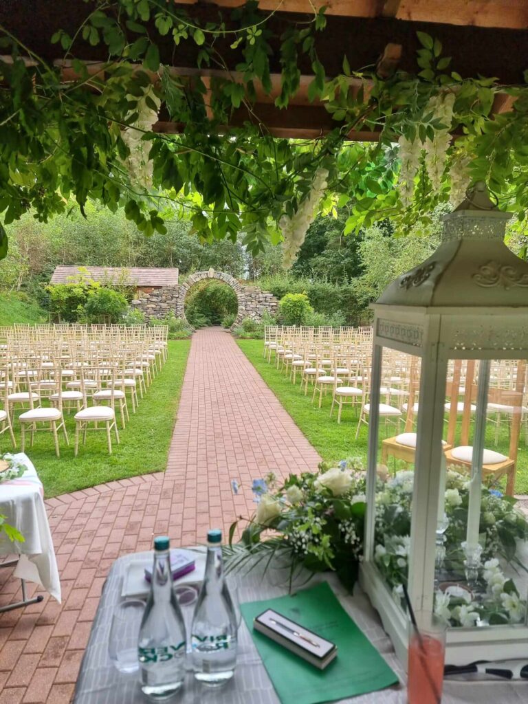 Outdoor wedding ceremony setup at Fernhill, featuring a stone archway, greenery, and white chairs arranged along a brick pathway.