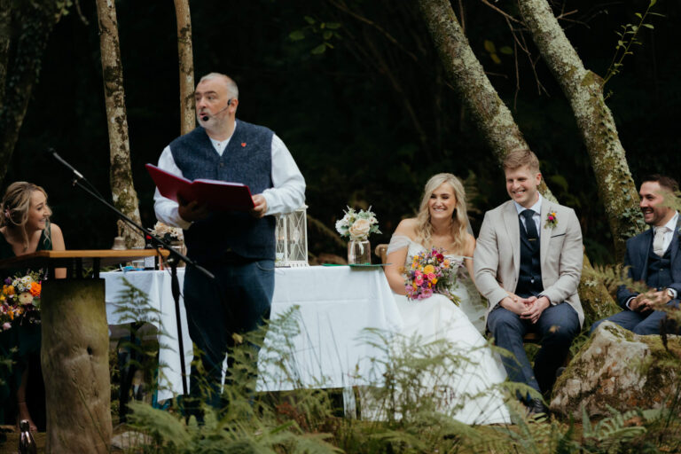 Wedding celebrant officiating an outdoor ceremony in a forest setting, with the bride, groom, and guests smiling in the background.