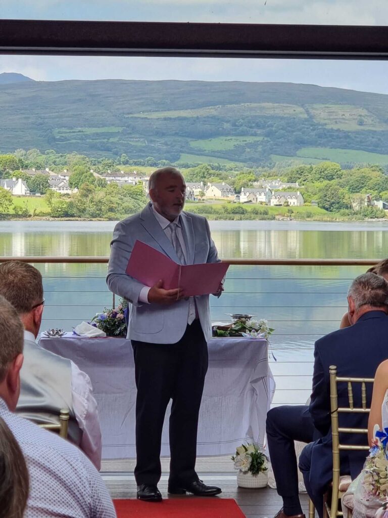 Wedding celebrant Brian Twomey officiating an outdoor ceremony at Sheen Falls with a scenic lake and mountains in the background.