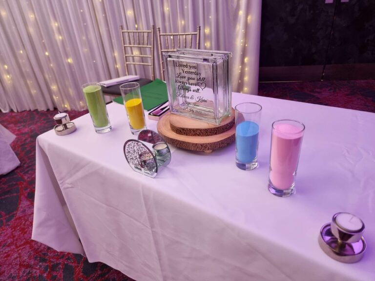 Unity sand ceremony table with colorful sand jars, a personalized glass vase, and candles, set against a backdrop of draped lights.