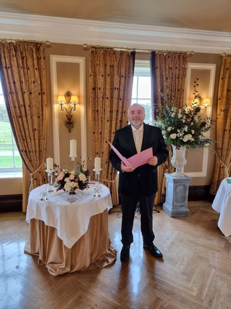 Wedding celebrant Brian in a formal black suit holding a pink folder, standing next to a beautifully decorated ceremony table with candles and flowers in Castlemartyr Hotel and Resort, an elegant venue.