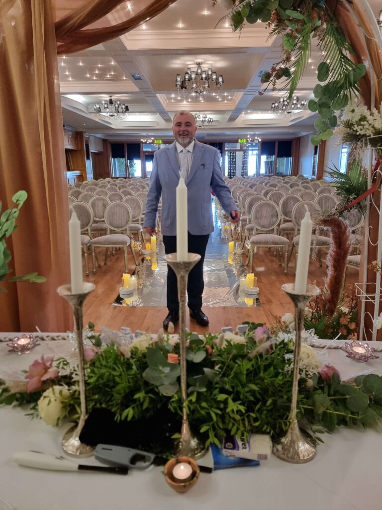 Wedding celebrant Brian Twomey in a light blue suit standing in an elegant ceremony room at The Garryvoe Hotel with floral decorations, candles, and a beautifully lit aisle.