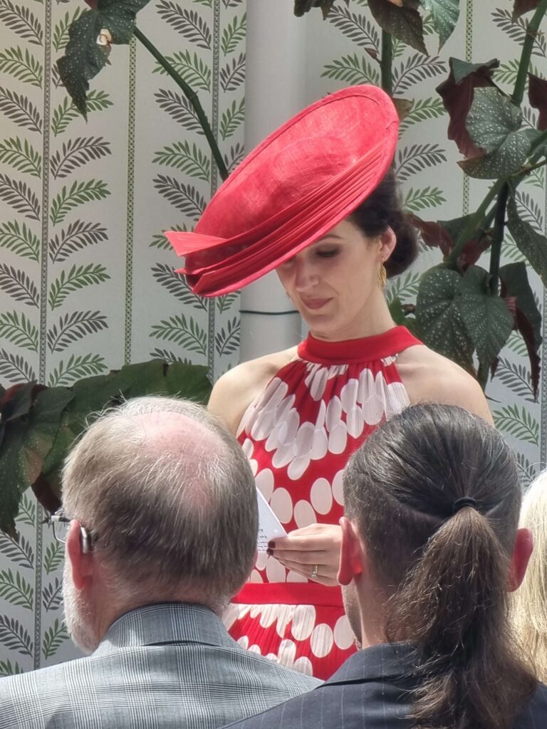 Wedding guest in a red polka-dot dress and a matching large red fascinator reading during a wedding ceremony at Ballymaloe.