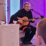 Musician John Spillane playing an acoustic guitar and singing during a wedding ceremony, with soft purple lighting in the background.